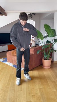 Jovem a fechar uma jaqueta cinza, em pé num chão de madeira numa sala de estar com um sofá laranja e uma grande planta verde em vaso.