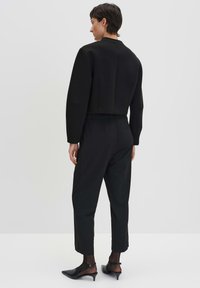 Veste noire courte avec un col rond et des épaules structurées, associée à un pantalon noir taille haute et des talons aiguilles noirs.