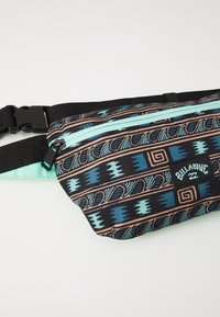 Sac banane en tissu noir avec des motifs tribaux en bleu et pêche. Doté d'une fermeture éclair vert menthe et d'une sangle noire réglable. Logo Billabong à l'avant.