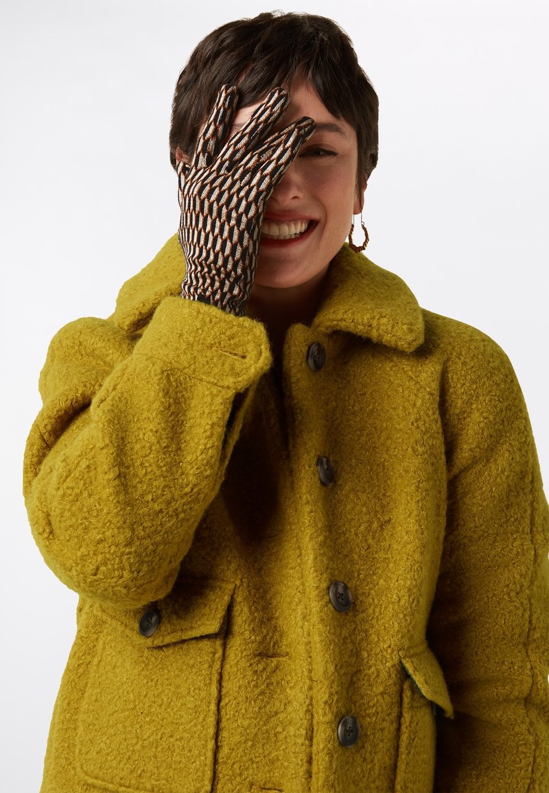Manteau en laine texturé de couleur jaune vif avec un col et des boutons à l'avant. La main montre un gant marron et crème à motifs, couvrant la moitié du visage.