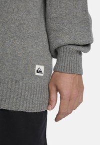 Grauer Strickpullover mit geripptem Saum und Bündchen, versehen mit mehrfarbigen Sprenkeln. Das Etikett mit dem Logo ist in der Nähe des Saums sichtbar. Eine Hand ruht dagegen.