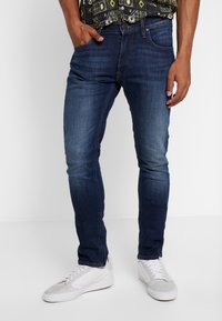 Mörkblå denimjeans med slim fit, med ton-i-ton sömmar, femficksdesign och slitningar på låren. Vita sneakers synliga.