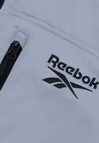 Jasnoszara sportowa kurtka z kieszenią na zamek. Ozdobiona czarnym logo "Reebok" i akcentami, wykonana z gładkiego, teksturowanego materiału.