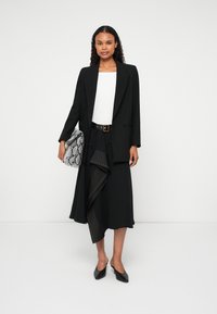 Melnā žakete virs baltā topa, kombinēta ar melnu svārku ar drapējuma dizainu un studētām akcentiem. Tur rokassomu ar čūskas rakstu.