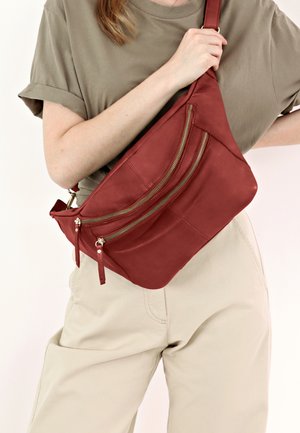 Red leather crossbody bag with three zippered pockets, a smooth texture, angled shape, and adjustable strap, worn against a light background.