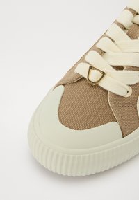 Baskets en toile brunes avec des lacets crème, un dessus texturé et une semelle en caoutchouc vert clair présentant des stries. Accent doré sur le laçage.