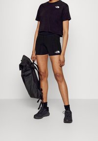 Černé zkrácené tričko a šortky, oboje se znakem The North Face. Černé tenisky a strukturovaný černý batoh držený v jedné ruce.