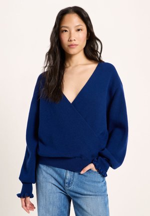 Frau mit langen dunklen Haaren trägt einen dunkelblauen Pullover mit tiefem V-Ausschnitt und hellblaue Jeans, steht mit einer Hand in der Tasche vor einem schlichten Hintergrund.