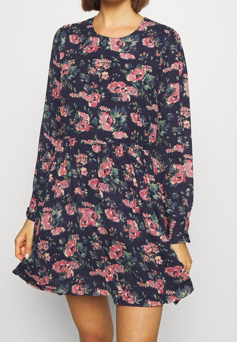 Tumma laivastonsininen kukallinen mekko, jossa pitkät hihat, vaaleanpunaiset ja vihreät kukkakuviot, kerrostettu vyötärö ja levenevä helma, kevyt kangas.