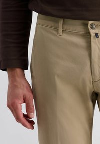 Beige Hose aus leichtem Stoff mit glatter Textur. Mit Knopfverschluss, Gürtelschlaufen undSeitentaschen. Hand ruht auf dem Stoff.