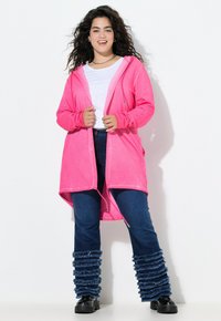 Cardigan rosa con cappuccio realizzato in tessuto morbido, caratterizzato da un design lungo e aperto. Abbinato a jeans blu scuro e gambali arricciati blu.