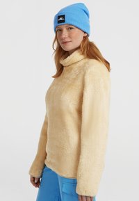 Lichtbeige fleece pullover met een hoge kraag en lange mouwen, met een zachte textuur. Gecombineerd met een felblauwe gebreide beanie.