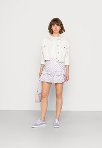 Jaqueta de ganga branca por cima de uma blusa branca, combinada com uma saia lilás com volantes e padrão floral. Completa com ténis lilás e uma pequena bolsa.