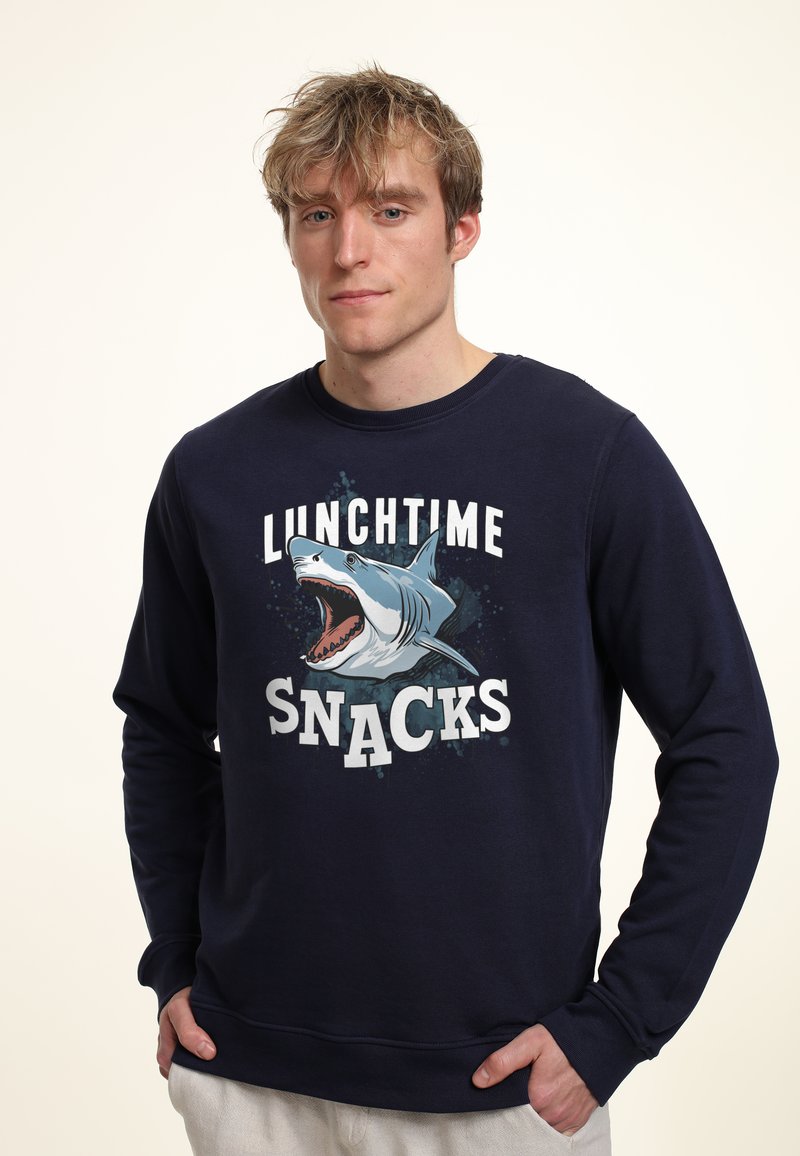 Sudadera de color azul marino con un gráfico de la boca de un tiburón y el texto "SNACKS EN LA HORA DEL ALMUERZO" en letras blancas y en negrita. Tela suave con mangas largas.