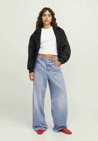 Svart kort bomberjacka över en vit tajt topp, kombinerad med ljusblå vida jeans och röda klackskor. Slät textur, avslappnad stil.
