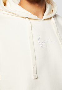 Světle béžová mikina s kapucí, vyrobená z měkké tkaniny, s šňůrkovou kapucí, přední kapsou a vyšitým logem na hrudi.