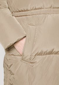 Veste matelassée beige avec une main visible glissée dans la poche latérale, montrant des coutures détaillées et une texture de tissu lisse.