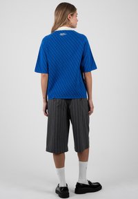 Mujer de pie, vistiendo una camisa de manga corta azul con estampado, pantalones cortos grises hasta la rodilla con rayas finas, calcetines blancos y mocasines negros sobre un fondo blanco.