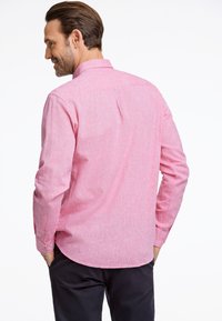 Vaaleanpunainen pitkähihainen paita, jossa on teksturoitu kangas, klassinen kaulus ja napilliset hihansuut. Takana on pleat lisäämään liikkuvuutta.