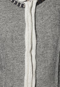Suéter gris con un diseño de punto texturizado, un acento central de cordón blanco y un detalle metálico en el collar.