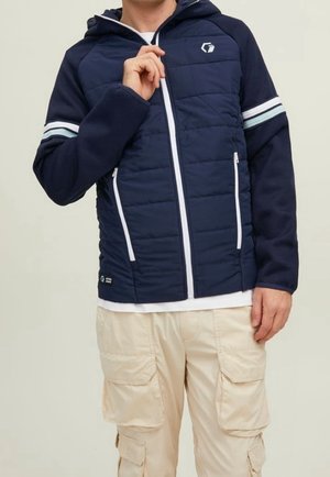 Homme portant une veste à capuche bleu marine avec une fermeture éclair blanche et des bandes sur les manches, ainsi qu'un pantalon cargo beige.