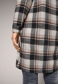 Camicia a quadri con motivi neri, bianchi e ruggine, realizzata in tessuto flanella. Presenta polsini con bottoni e un orlo leggermente curvato.