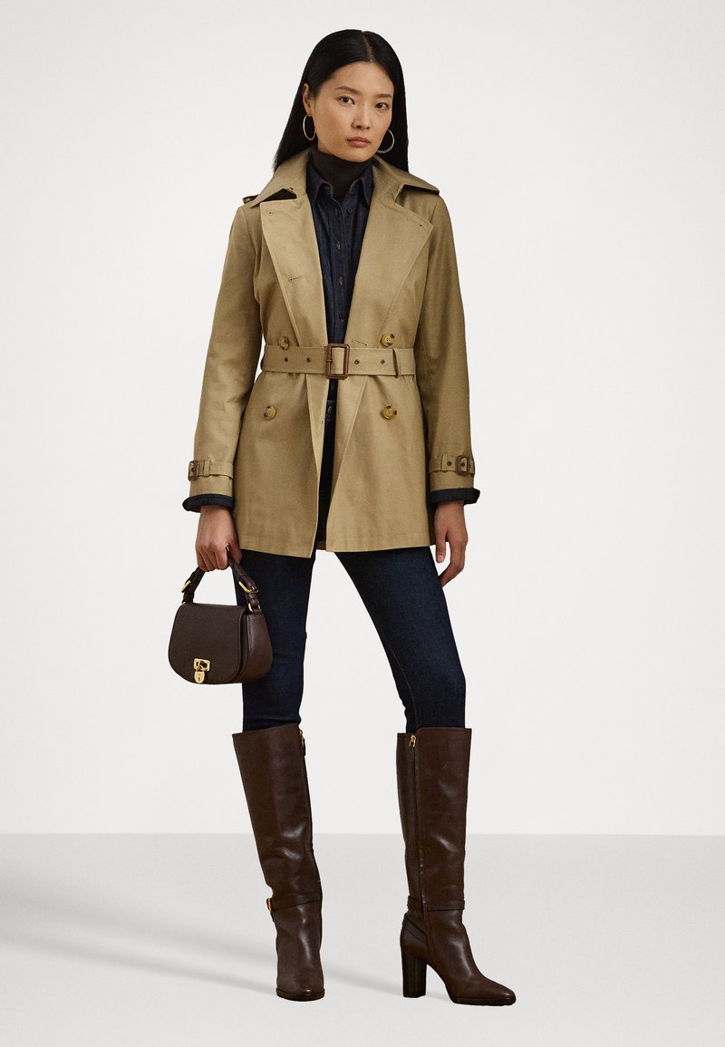 Beige trench coat with double-breasted buttons, belt detail, and side pockets. Worn over a buttoned navy shirt and paired with dark jeans and brown knee-high boots.