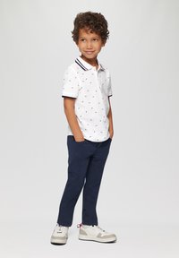 Witte polo met marineblauwe kraag en een patroon van strikjes, gecombineerd met marineblauwe broek en grijze sneakers, staand met de handen in de zakken.