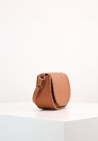Borsa a tracolla in pelle color tan con una forma curva, cuciture dettagliate lungo il bordo e una tracolla regolabile. Texture liscia.