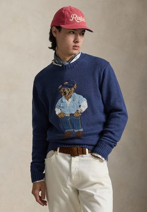 Tengerészkék kötött pulóver, amely egy medve figurát ábrázol farmer öltözékben, bordázott mandzsettákkal és szegéllyel, krémszínű nadrággal és piros sapkával párosítva.