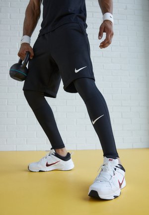 Personne en tenue de sport noire et chaussures blanches Nike tenant un kettlebell, debout sur un sol jaune contre un mur en briques blanches.