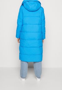 Lång, vadderad blå kappa med huva. Har horisontella quiltade sektioner och rundad fåll. Bärs över ljusblå jeans och vita sneakers.