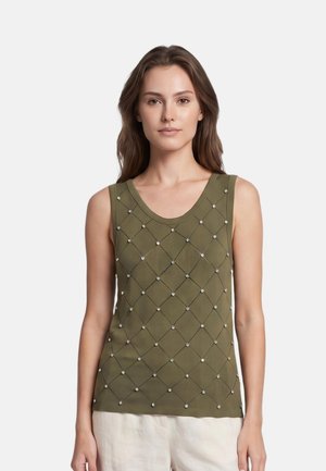 Jonge vrouw met lang bruin haar die een olijfgroene mouwloze top draagt met een diamantpatroon en kleine witte kraaltjes, gecombineerd met lichte broek.