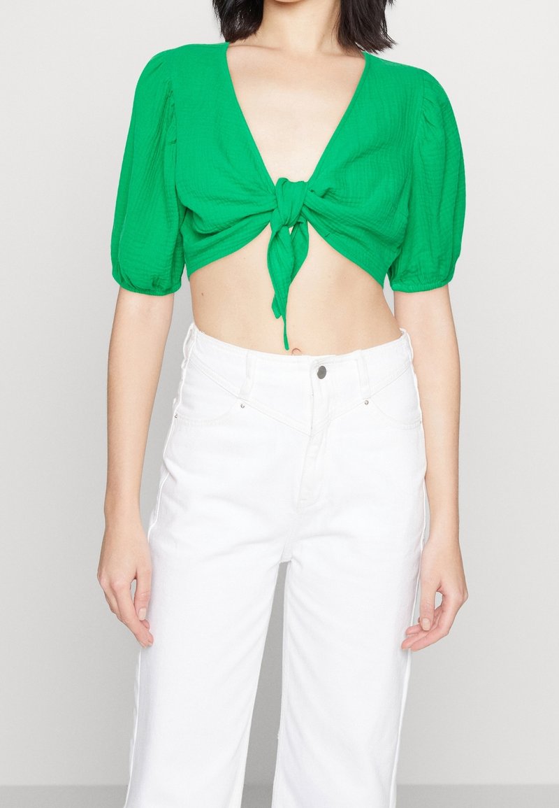 Blouse verte raccourcie avec un col en V profond et un lien à l'avant. Les manches bouffantes mettent en valeur le design. Associée à un pantalon blanc taille haute.