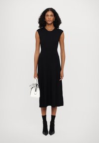 Vestido preto de malha canelada com corpete justo e saia rodada. Usado com meias pretas até ao tornozelo e sapatos pretos de ponta. Segurando uma pequena mala branca.