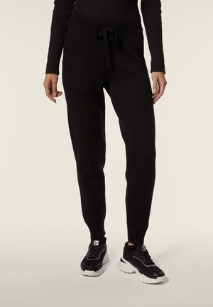 Pantaloni sportivi neri con una texture morbida, vita elastica e cordino. Presentano tasche anteriori e polsini affusolati. Indossati con sneakers nere.