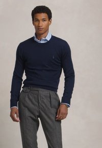 Pull en laine marine à col rond porté par-dessus une chemise à col bleu clair, associé à un pantalon gris à plis. Texture et coupe du tissu notables.
