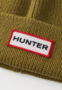 Maslinasto zelena pletena kapa s rebrastom teksturom, koja ima istaknut bijeli pečat s crvenom bordurom i crnim natpisom "HUNTER."
