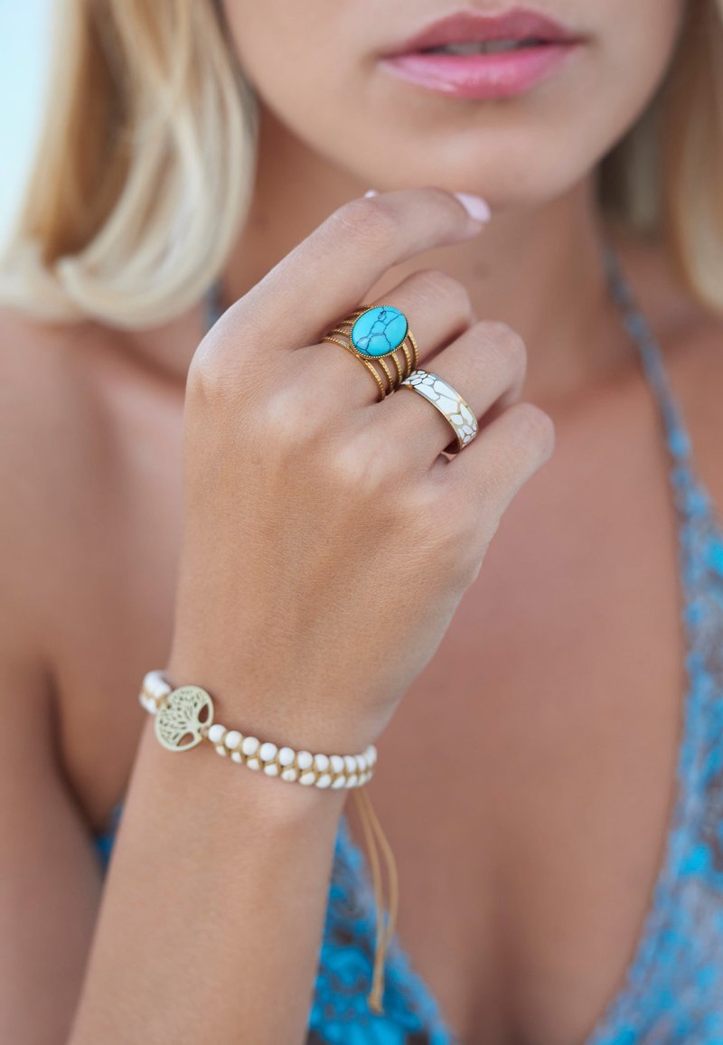 Gouden ringen met een turquoise steen, gecombineerd met een gestructureerde witte band. Een armband met witte kralen bevat een levensboombedel.