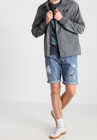 Chaqueta ligera gris sobre una camiseta azul verdosa, combinada con pantalones cortos de mezclilla desgastados y zapatillas ligeras. El conjunto presenta un estilo casual y relajado.