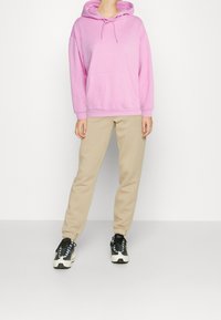 Felpa oversize rosa con tasca frontale e cordoncino. Pantaloni della tuta beige. Sneakers bianco e nere con lacci blu.