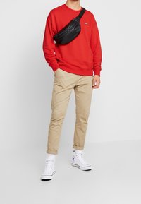 Uomo in felpa rossa, pantaloni beige arrotolati, sneakers alte bianche e borsa a tracolla nera sul torace, davanti a uno sfondo bianco.