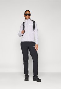 Šviesiai purpurinė fliso zip-up striukė, juodos kelnes su kišenėmis, juodas kuprinė ir tamsūs batai. Modelis stovi su rankomis ant klubų.