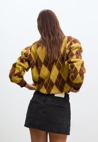 Femme aux longs cheveux bruns portant un pull jaune et marron à motif losanges et une mini-jupe en jean noire, debout les mains sur les hanches, de dos.