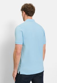 Man die met de rug naar de camera staat, gekleed in een lichtblauw poloshirt met korte mouwen en een donkere spijkerbroek, tegen een effen witte achtergrond.