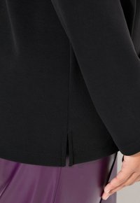 Top negro de manga larga con cuello redondo, aberturas laterales en el dobladillo y tela suave texturizada, combinado con pantalones morados brillantes.