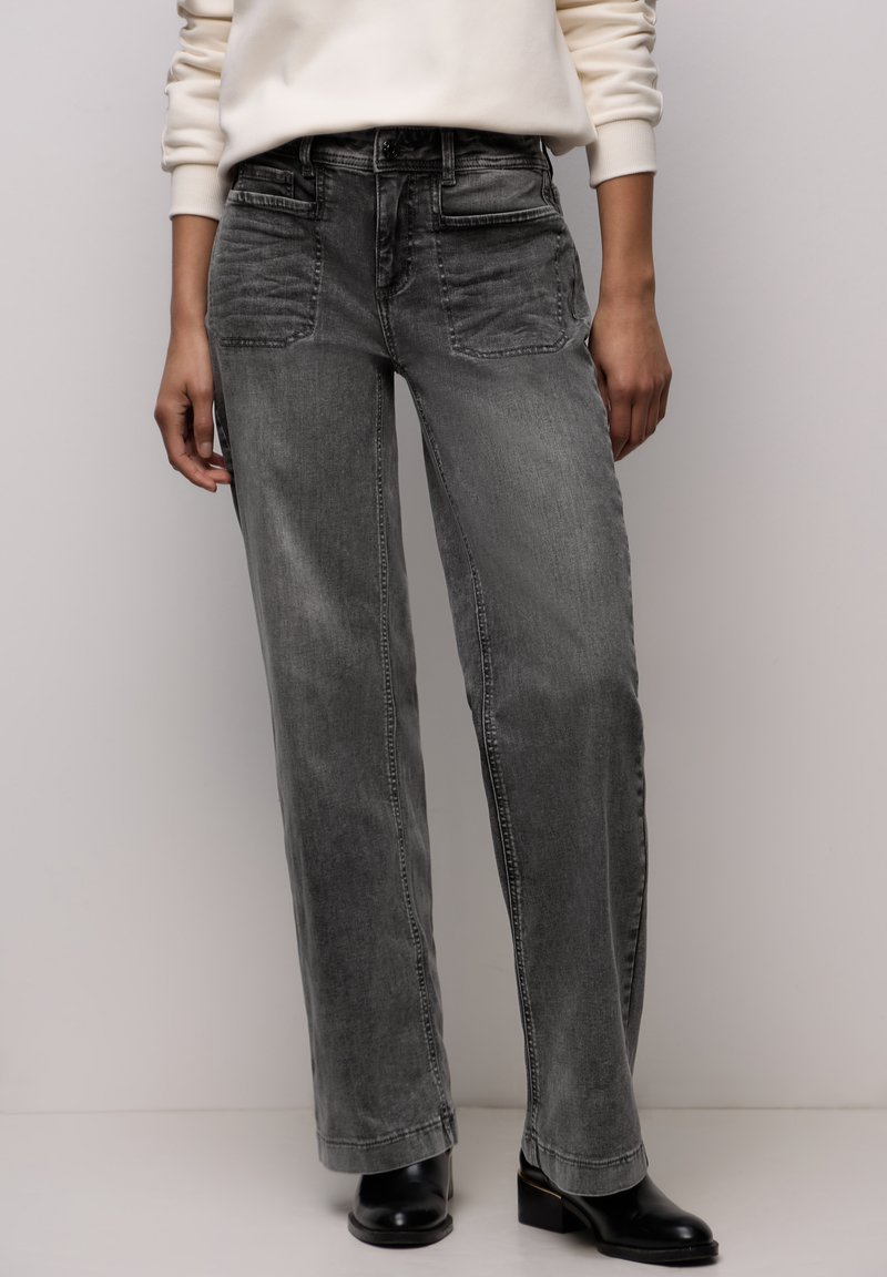 Jean à jambes larges en denim gris clair, avec poches avant, une texture subtilement délavée, et des détails de couture latérale, associé à des bottines noires.