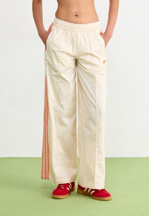 Femme portant un pantalon large crème taille haute avec des rayures orange et un logo, associé à des baskets rouges et blanches, debout sur un sol vert.