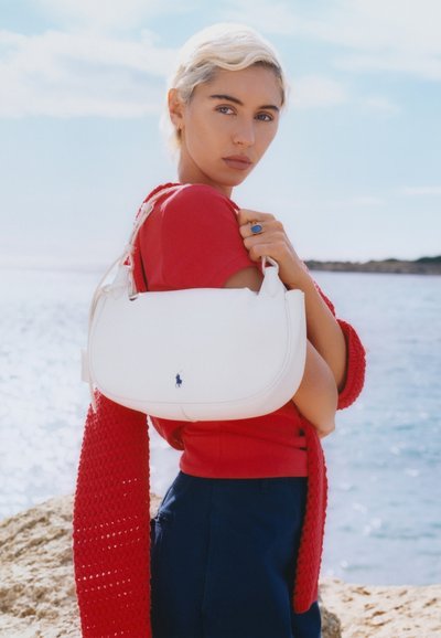 Femme aux cheveux blonds courts, vêtue d'un top rouge et d'une écharpe, tenant un sac à bandoulière blanc, debout près d'une rive rocheuse ensoleillée avec une eau calme en arrière-plan.