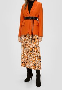 Kvinna iklädd orange kavaj med svart bälte, blommig plisserad kjol, svart polokrage och svarta klackboots som står framför en enfärgad bakgrund.